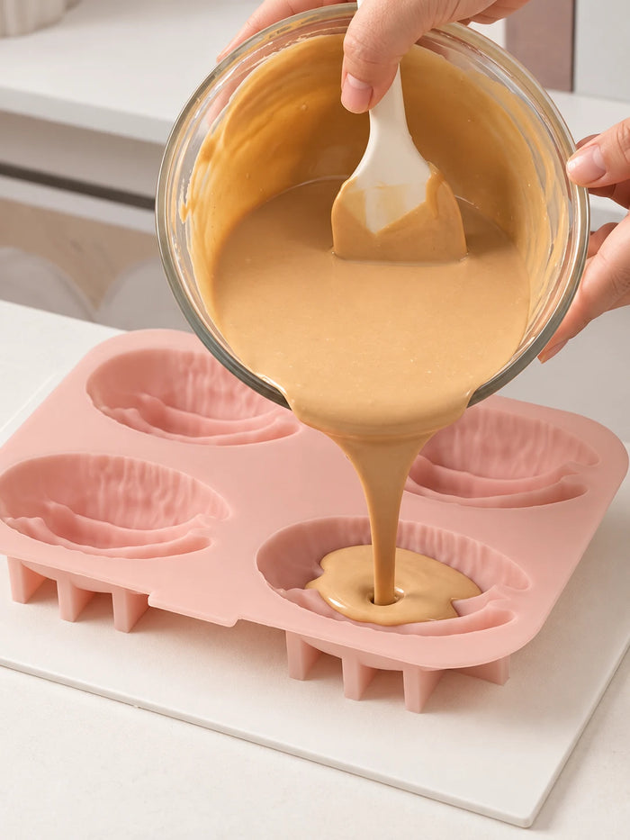 Tan mixture pours from a glass bowl into one of four textured pecan kernel cavities in a pink mold, with a white spatula held inside the bowl.