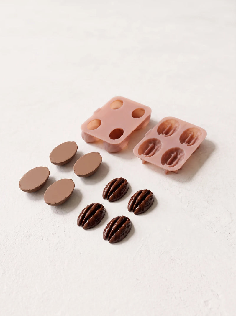 Two pink molds behind separate cake components: four smooth tan oval bases and four glossy brown pieces shaped like pecan kernels.