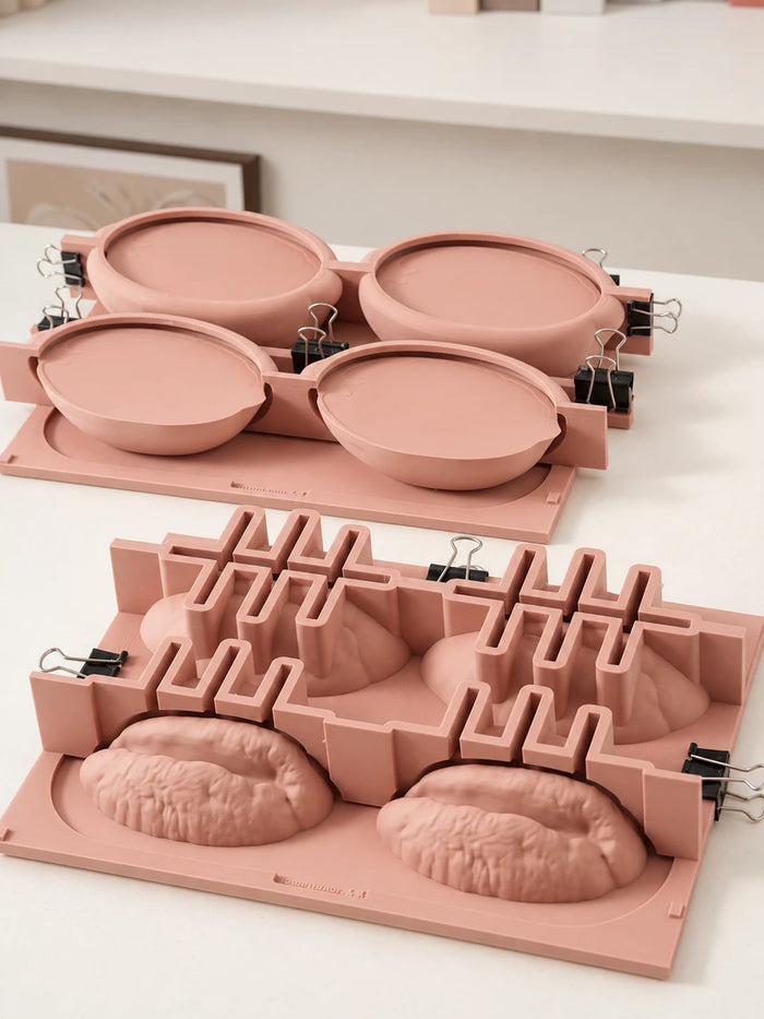 Two pink master-model plates show oval shell forms at the back and ridged pecan kernel forms in front, with black clips securing the surrounding frames.