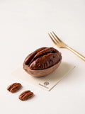 A pecan-shaped cake on an ivory card, showing a glossy dark brown kernel top and matte tan shell base, with loose pecans and a gold-colored fork nearby.