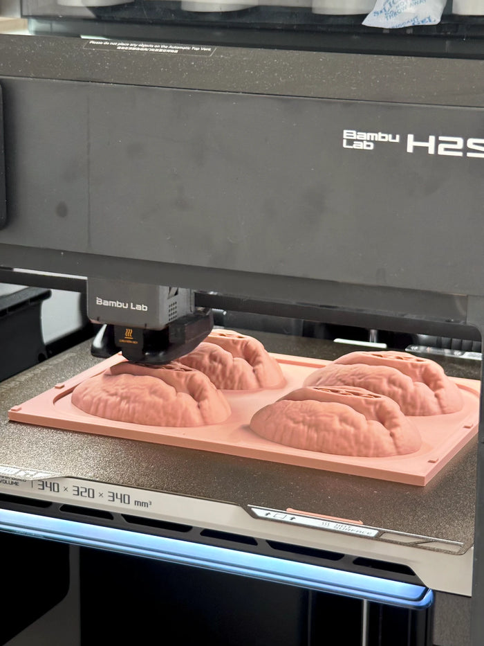 A 3D printer head sits above a pink plate carrying four pecan kernel master models with deeply grooved surfaces.