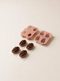 Four pecan-shaped cakes with glossy brown kernel forms and tan shell-shaped bases, arranged on individual ivory cards in front of two pink molds.