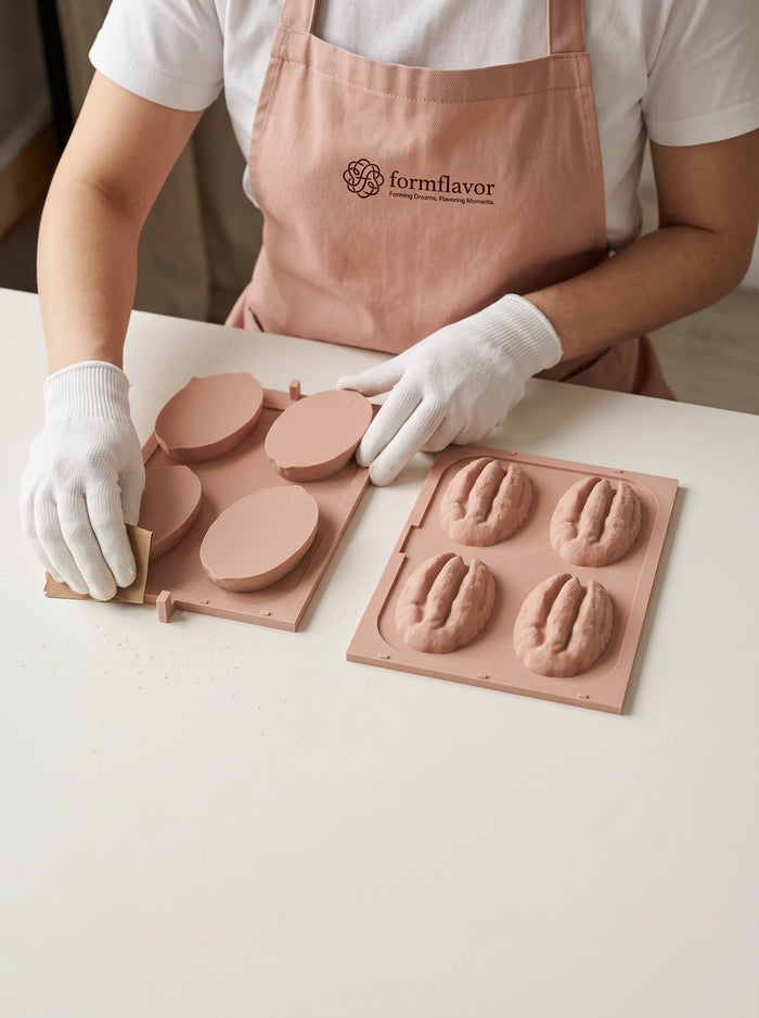 Pink pecan shell and kernel master models on two plates, with a gloved hand holding sandpaper against the shell plate's edge.